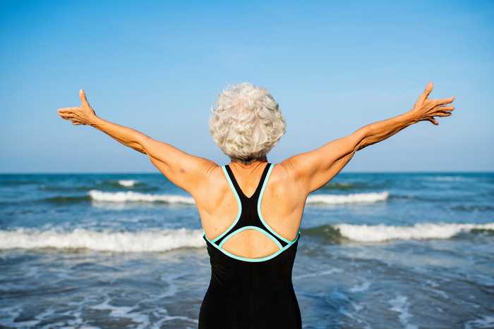 An older lady with grey curly hair in a cross back black swimming costume facing away from the camera, arms raised towards the waves